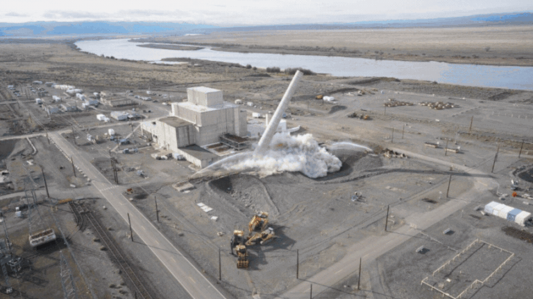 Aerial view of demolition work at the Hanford Site near the Columbia River, with a tall stack collapsing beside an industrial facility.
